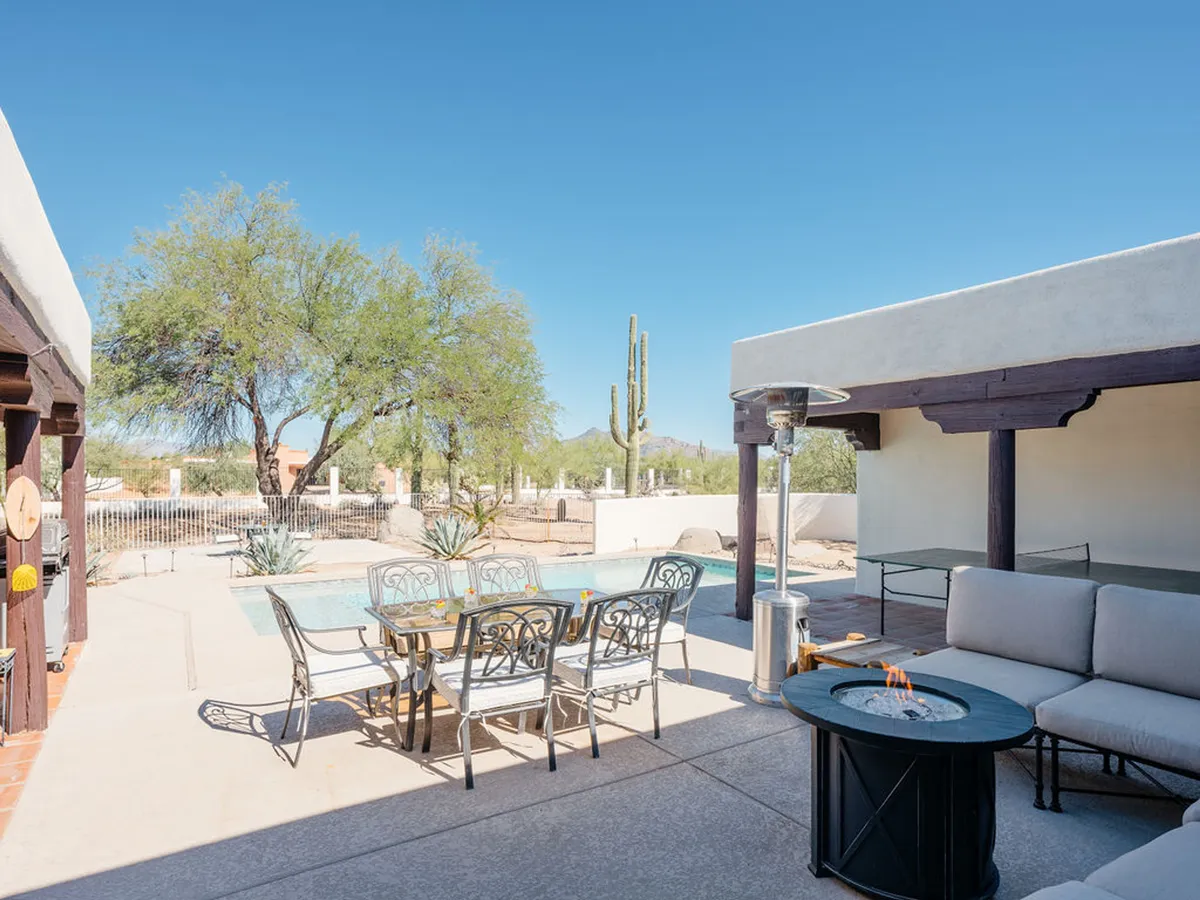 A tranquil backyard scene highlights a crystal-clear swimming pool, ample outdoor lounge seating, a fire pit area, and signature desert landscaping with cacti in the background. The arrangement feels both luxurious and inviting, reflecting a perfect space for sunbathing, socializing, or enjoying quiet moments by the pool or fire.