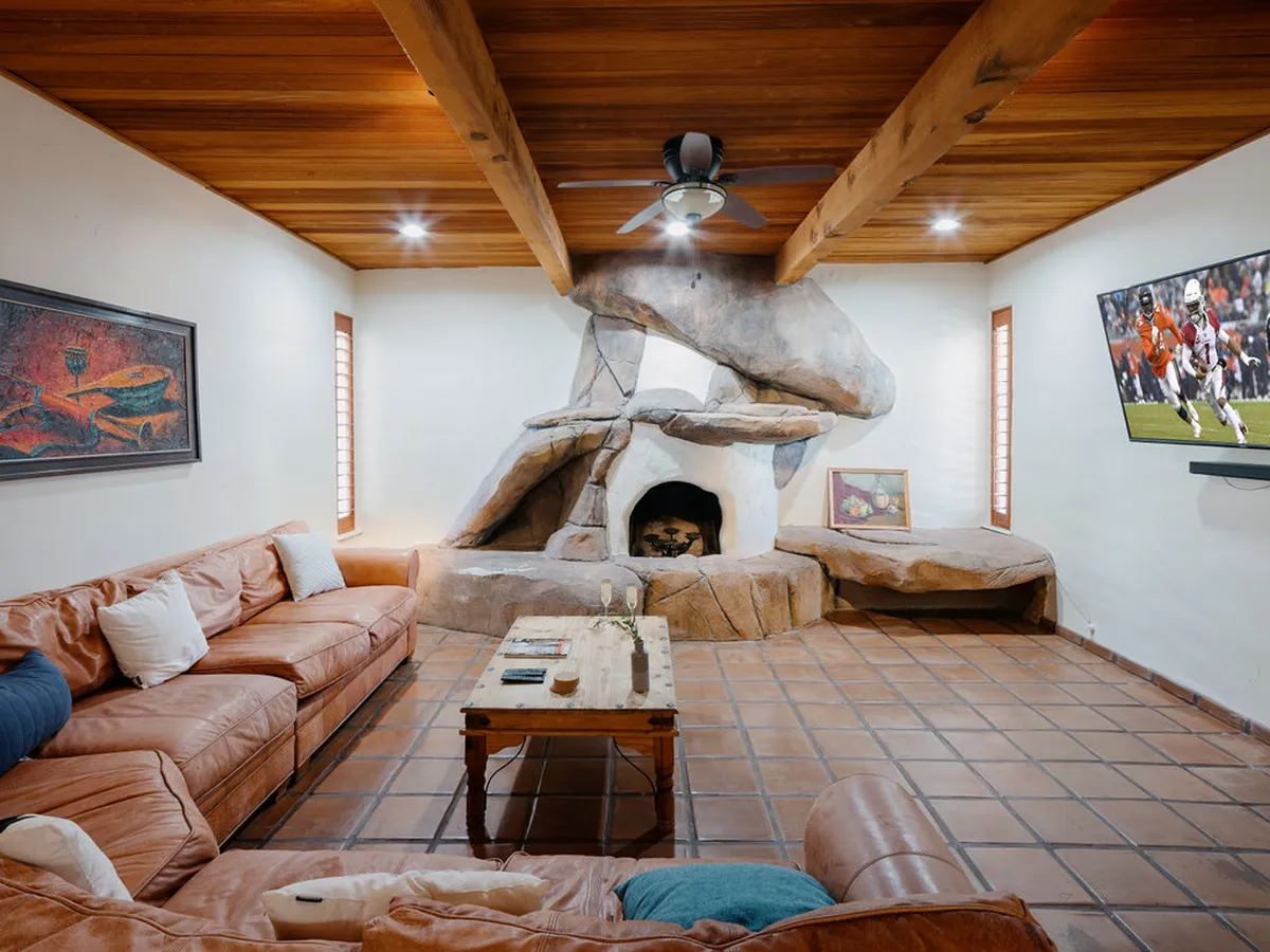 A spacious living room featuring a large stone fireplace as the central focal point, complemented by a comfortable leather sectional sofa, wooden coffee table, mounted television, and artwork on the wall. The space exudes a relaxed and homey mood, perfect for gatherings, movie nights, and cozy conversations.