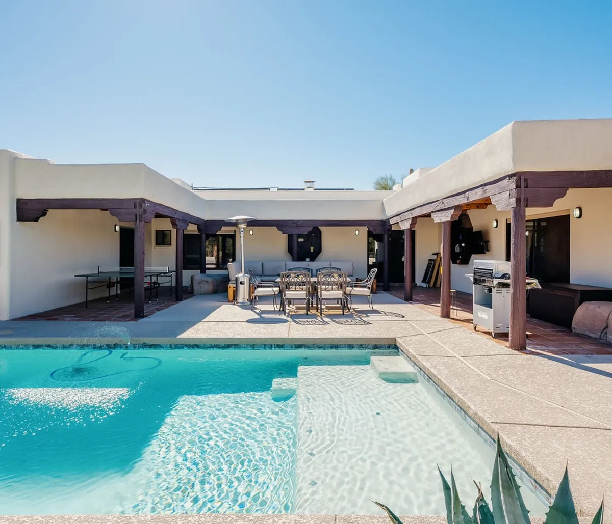 The image showcases a stunning backyard oasis, featuring a sparkling swimming pool surrounded by a spacious patio with an inviting outdoor dining area. A built-in grill hints at the promise of delicious barbecues, while plush lounge seating offers a perfect spot to relax or socialize under the Arizona sun. The mood is leisurely and social, highlighting the ideal setting for poolside gatherings and sunset dinners with family and friends.