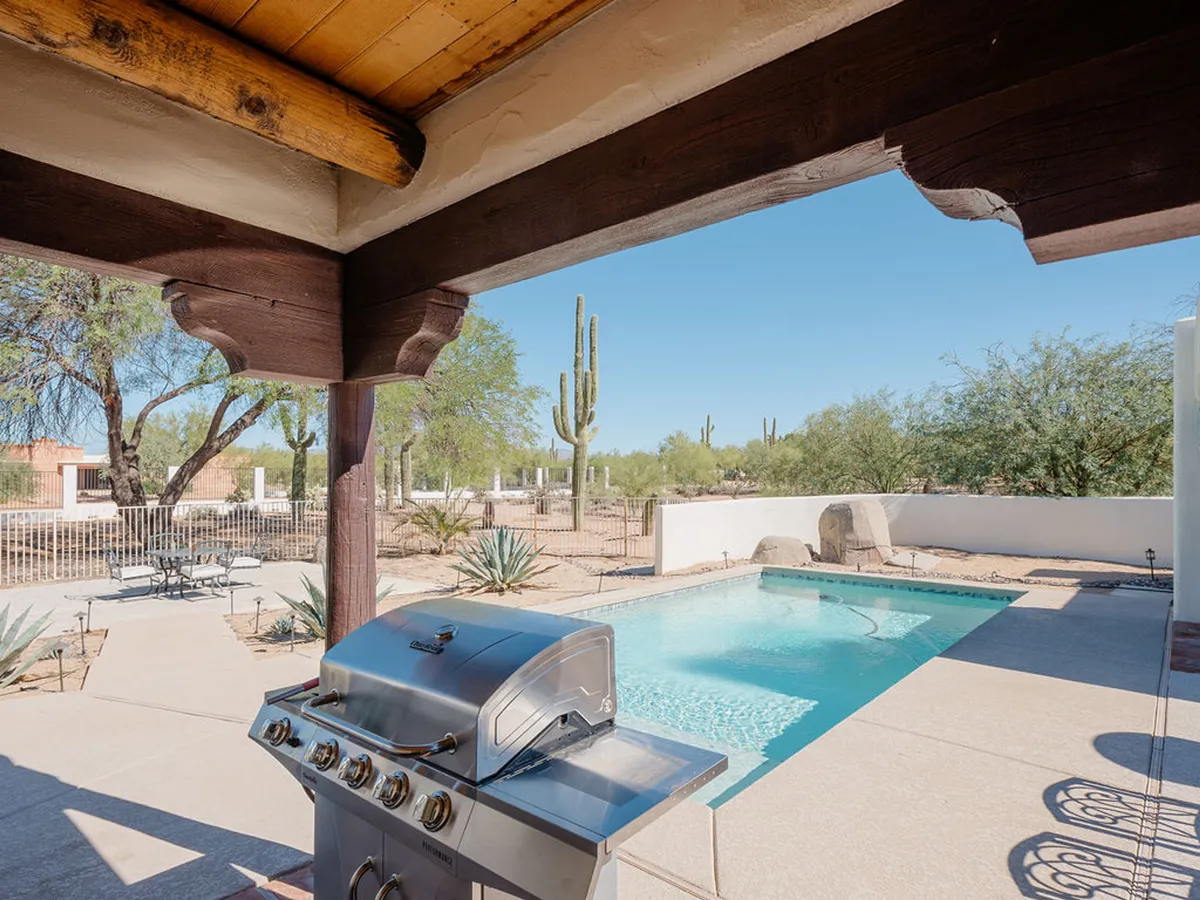 A sparkling swimming pool set against a desert landscape, featuring a grill for outdoor cooking, surrounded by cacti and seating areas. The space is designed for relaxation, social gatherings, and poolside barbecues.