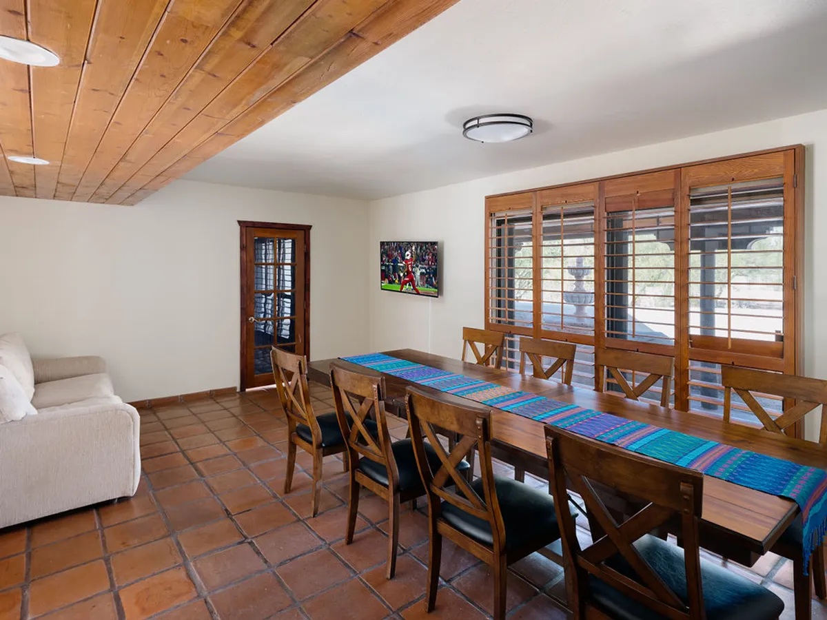 A dining area featuring a long table set up for group meals, a TV on the wall, and a sofa nearby. The environment feels comfortable and sociable, ready for family dinners, celebrations, or casual gatherings.