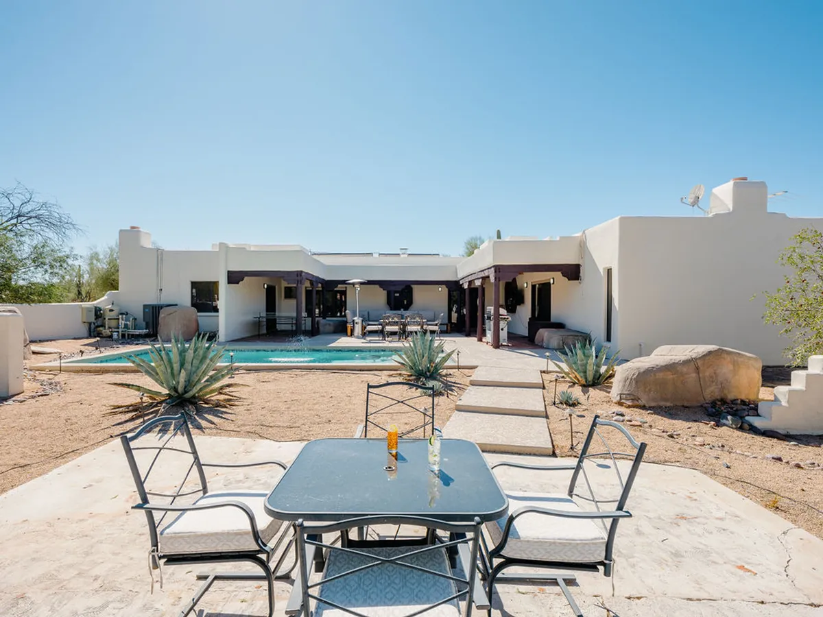 An elegant patio scene features a sun-kissed swimming pool, a spacious outdoor dining table ideal for barbecues, and the surrounding desert landscaping with agave plants. Comfortable patio furniture invites relaxation, while the overall layout encourages seamless movement between swimming, dining, and lounging.