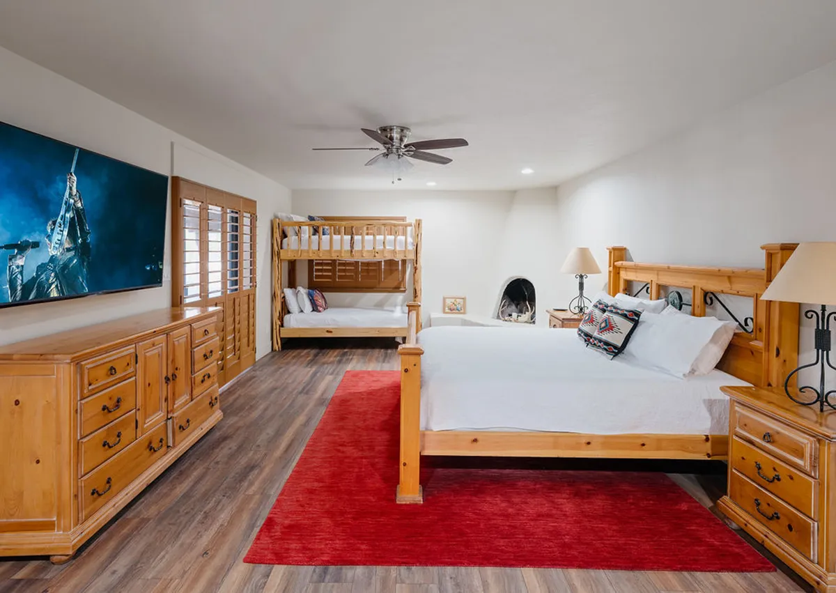 A warm and inviting bedroom showcasing a large bed, an adjacent bunk bed, a TV, a dresser, and a cozy rug underfoot. The arrangement suggests a shared but private space, ideal for family movie nights and relaxation after Arizona adventures.