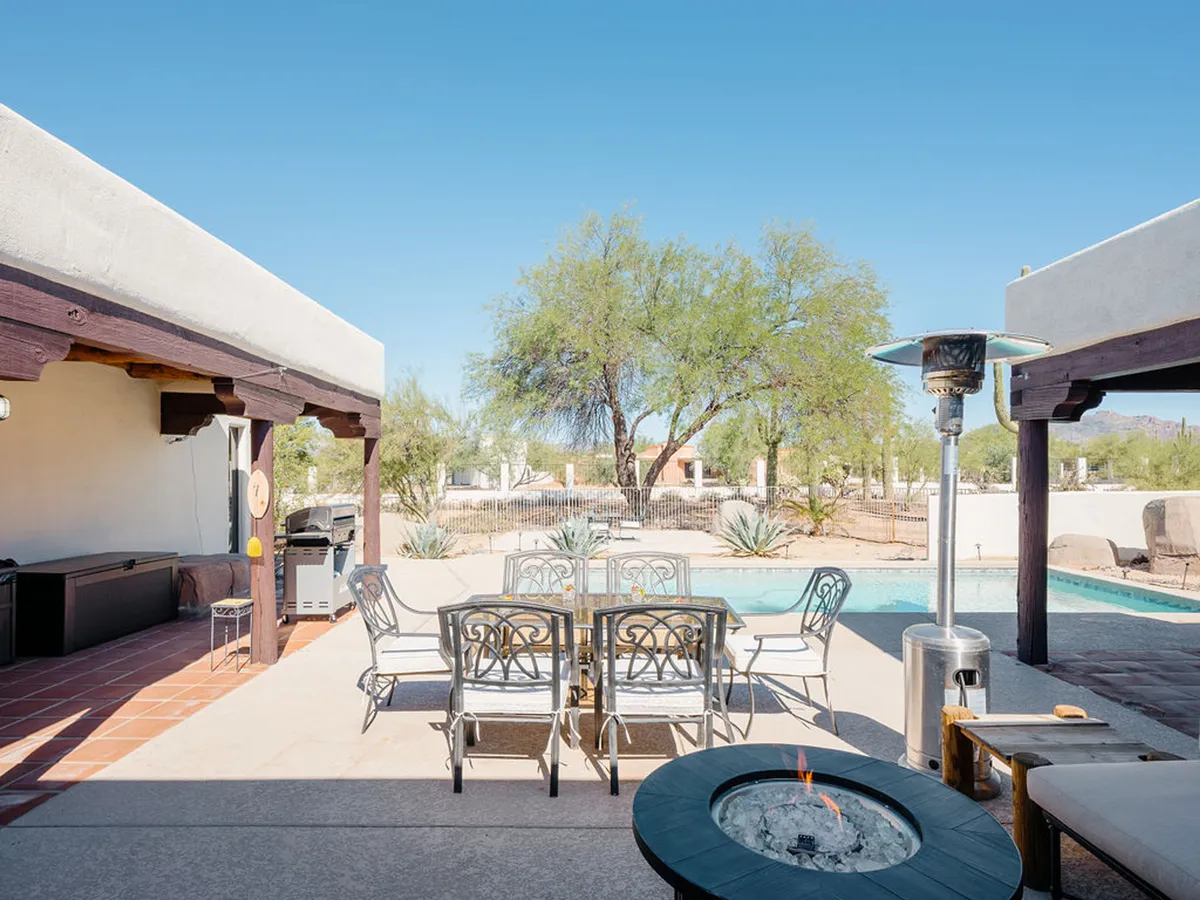 This exterior image features a beautifully arranged outdoor space, with a dining table set beside a sparkling swimming pool and a cozy fire pit. Lush greenery and patio seating frame the area, set under a vibrant, sunset-tinged sky that gives the space a warm, welcoming atmosphere. The setting captures multiple elements: pool, fire pit, outdoor dining, and a seamless transition between indoor comfort and outdoor enjoyment.